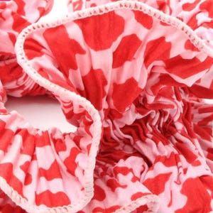 May include: Close-up of a red and pink patterned hair scrunchie. The scrunchie is made of fabric with a ruffled edge.