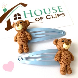May include: Two light blue alligator clips with brown teddy bear charms. The teddy bears have a textured fur design and black eyes.