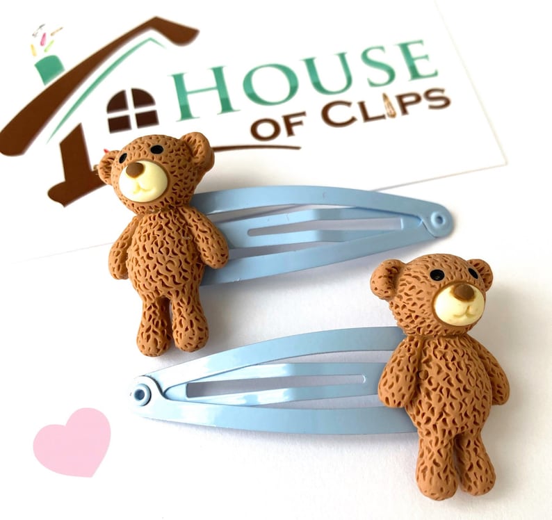 May include: Two light blue alligator clips with brown teddy bear charms. The teddy bears have a textured fur design and black eyes.