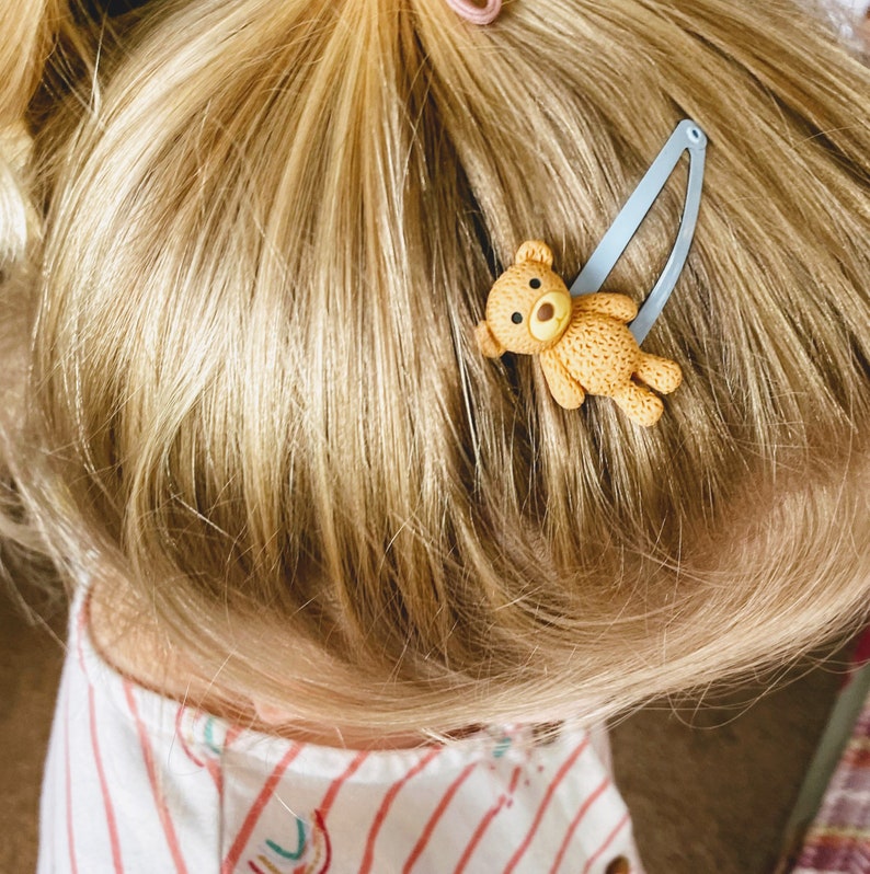 May include: A blue hair clip with a brown teddy bear charm. The teddy bear has a light brown nose and black eyes.