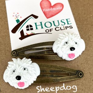 May include: Two bronze-colored barrettes with white sheepdog faces and pink tongues. The barrettes are next to a card with the text "HOUSE OF CLIPS" and a red heart that says "Handmade". The word "Sheepdog" is written below the barrettes.
