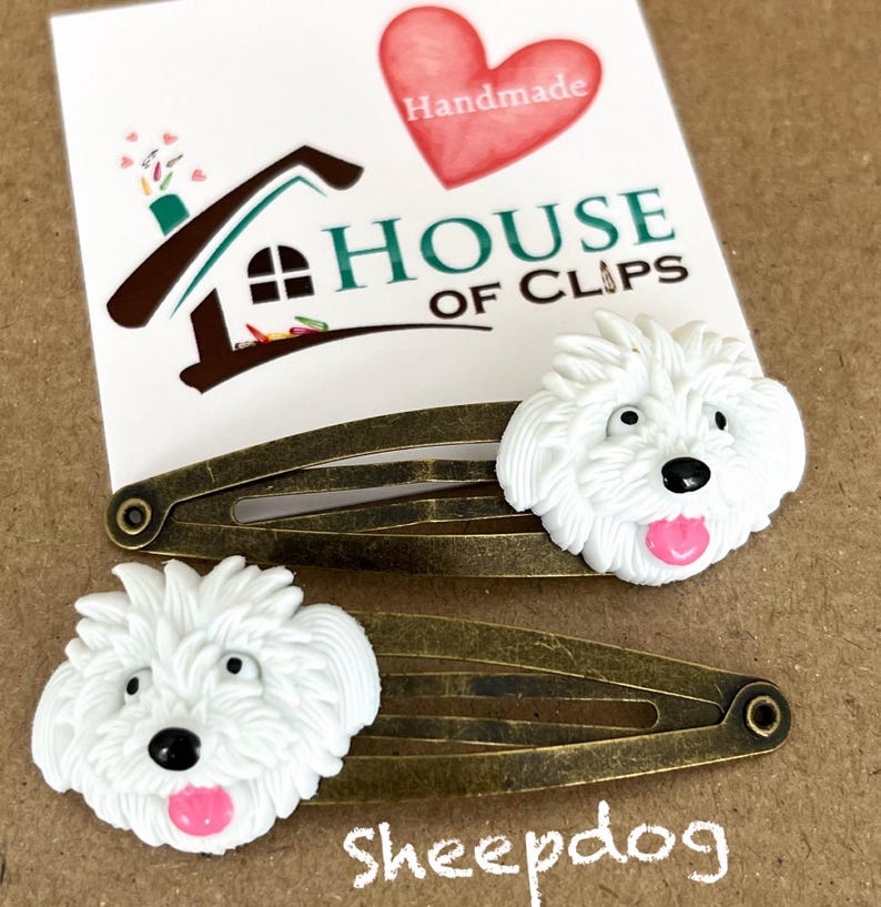 May include: Two bronze-colored barrettes with white sheepdog faces and pink tongues. The barrettes are next to a card with the text "HOUSE OF CLIPS" and a red heart that says "Handmade". The word "Sheepdog" is written below the barrettes.