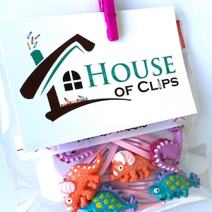 May include: A clear plastic bag containing hair clips with glittery dinosaur-shaped charms in orange, purple, and blue. A white card attached with a pink clip features the text House of Clips and a house-shaped logo with small clip icons.