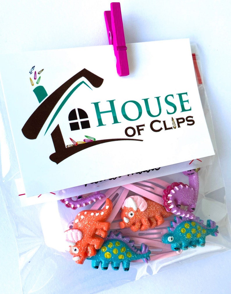 May include: A clear plastic bag containing hair clips with glittery dinosaur-shaped charms in orange, purple, and blue. A white card attached with a pink clip features the text House of Clips and a house-shaped logo with small clip icons.
