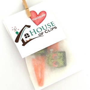Pode incluir: Um pequeno envelope branco com um cora&ccedil;&atilde;o vermelho e o texto "Handmade House of Clips" na frente. O envelope cont&eacute;m dois grampos de cabelo laranja e verde.