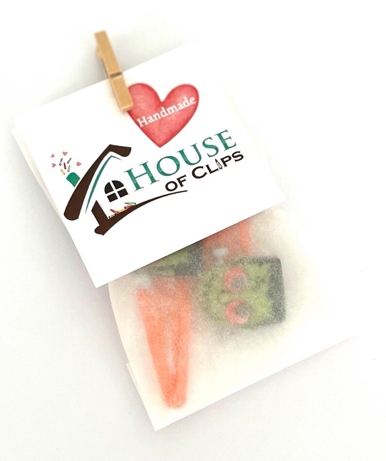 Pode incluir: Um pequeno envelope branco com um cora&ccedil;&atilde;o vermelho e o texto "Handmade House of Clips" na frente. O envelope cont&eacute;m dois grampos de cabelo laranja e verde.