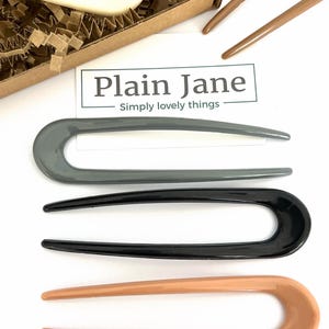 May include: Four hair pins in different colors: gray, black, beige, and white. The hair pins are all the same shape, with a long, thin, curved handle and a rounded end. The hair pins are arranged in a row, with the white hair pin at the top and the beige hair pin at the bottom. The text "Plain Jane - Simply lovely things" is visible in the background.