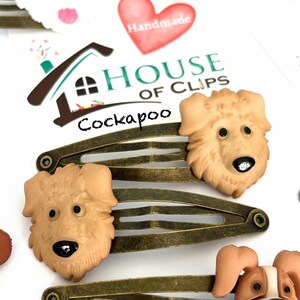 May include: Hair clips featuring Cockapoo dog faces. The clips are bronze-colored metal with a snap closure. The dog faces are tan with black noses and eyes. The image also includes a white dog face and a red heart with the word "Handmade".