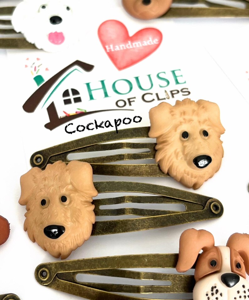 May include: Hair clips featuring Cockapoo dog faces. The clips are bronze-colored metal with a snap closure. The dog faces are tan with black noses and eyes. The image also includes a white dog face and a red heart with the word "Handmade".