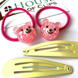 May include: Two pink bear hair ties with glitter and two yellow plastic hair clips. The hair ties are on a pink background with the text "House of Clips".