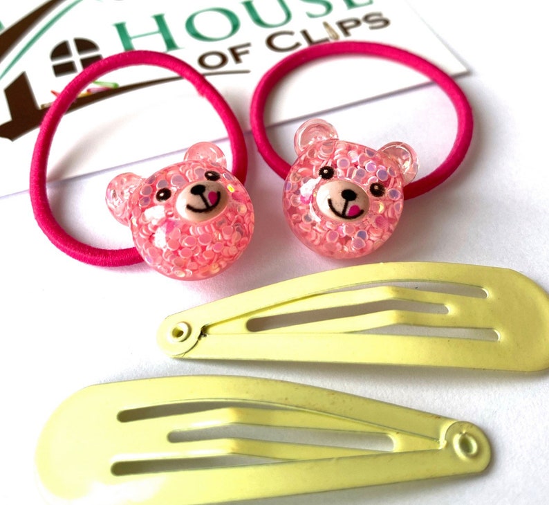 May include: Two pink bear hair ties with glitter and two yellow plastic hair clips. The hair ties are on a pink background with the text "House of Clips".