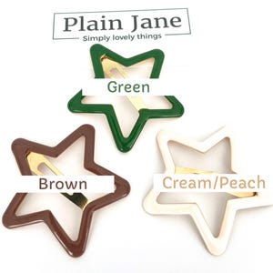 Pu&ograve; includere: Tre fermagli per capelli a forma di stella in verde, marrone e crema/pesca. Ogni fermaglio ha una base in metallo dorato. La parte superiore dell'immagine presenta il testo "Plain Jane" e "Simply lovely things".