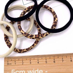 May include: A set of hair ties in black, beige, and leopard print. The hair ties are 5 cm wide and are shown on a wooden ruler.