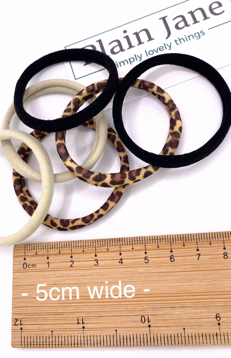 May include: A set of hair ties in black, beige, and leopard print. The hair ties are 5 cm wide and are shown on a wooden ruler.
