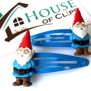 May include: Two blue plastic hair clips with a gnome charm on each. The gnomes are wearing red hats, blue shirts, brown pants, and white beards.