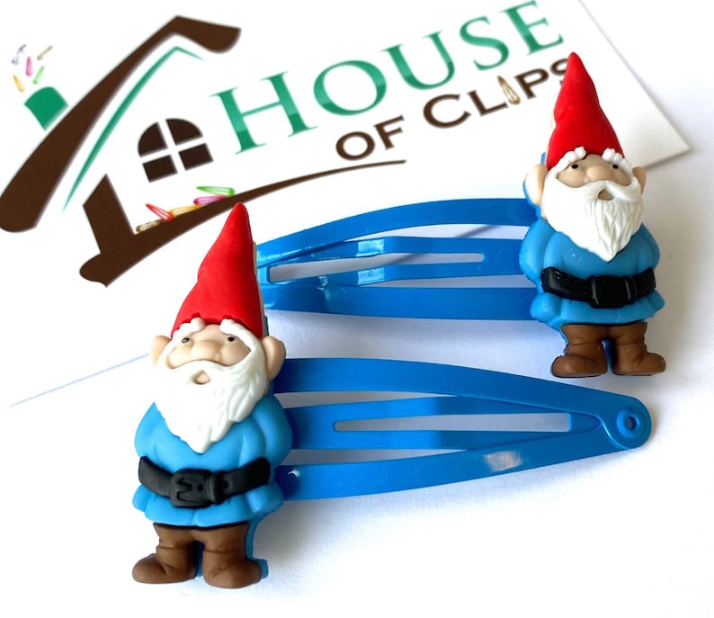 May include: Two blue plastic hair clips with a gnome charm on each. The gnomes are wearing red hats, blue shirts, brown pants, and white beards.