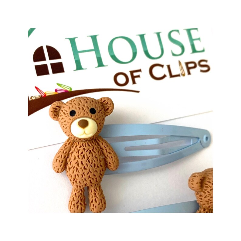 May include: A set of two blue hair clips with brown teddy bear charms. The teddy bears have black eyes, a brown nose, and a white mouth.