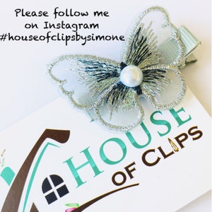 May include: A silver and black butterfly hair clip with a pearl accent. The clip is on a white card with the text "HOUSE OF CLIPS" and the hashtag #houseofclipsbysimone.