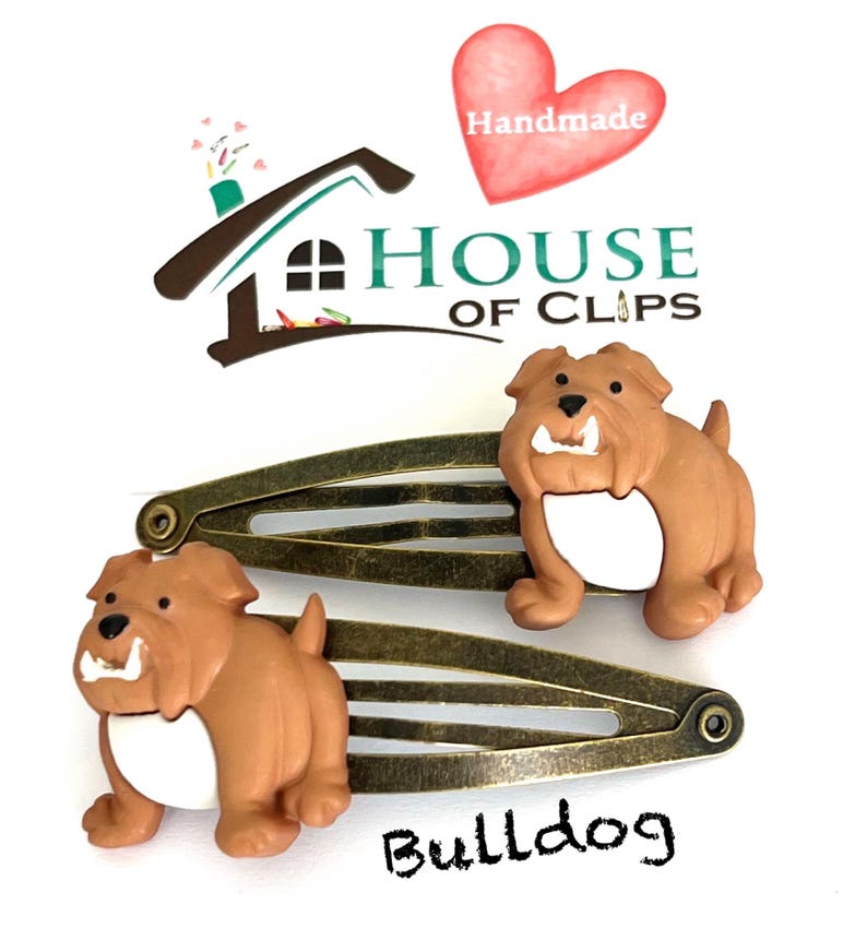 May include: Two bronze-toned metal hair clips, each adorned with a brown bulldog figurine with a white belly. The clips are set against a white background, with the word "Bulldog" and the "House of Clips" logo visible.