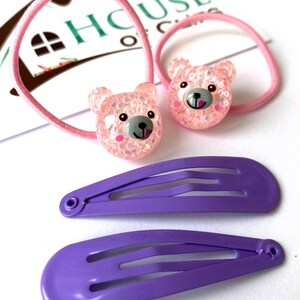 May include: Set of hair accessories featuring two pink rubber bands with sparkly pink bear charms and two purple plastic barrettes.