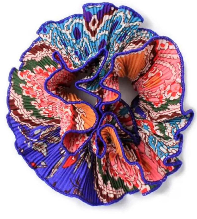 May include: A colorful hair scrunchie with a blue, orange, pink, and green paisley pattern. The scrunchie is made of pleated fabric and has a blue trim.