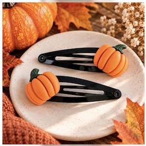 May include: Two black metal snap hair clips, each featuring a three-dimensional orange pumpkin charm with a small green stem attached to the top. The clips are positioned on a light-coloured, speckled surface, showcasing the pumpkin details.