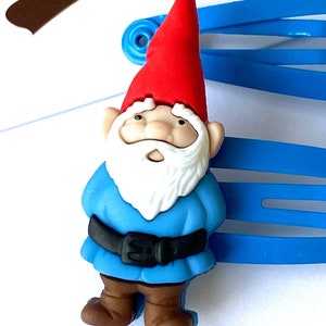May include: A blue plastic hair clip with a small gnome figurine attached. The gnome is wearing a red hat, a blue shirt, a black belt, and brown shoes.