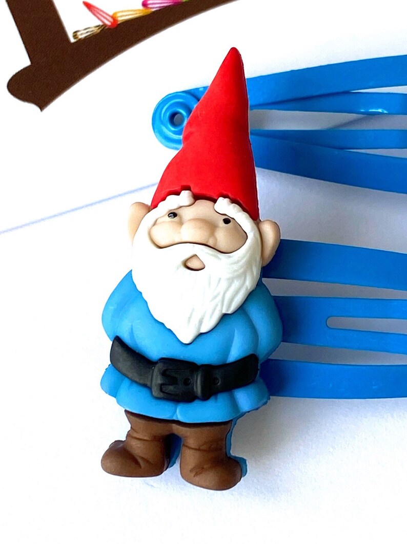 May include: A blue plastic hair clip with a small gnome figurine attached. The gnome is wearing a red hat, a blue shirt, a black belt, and brown shoes.