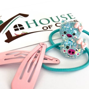 May include: Two turquoise hair ties with a blue pig face charm and a pink bow. Two pink hair clips.