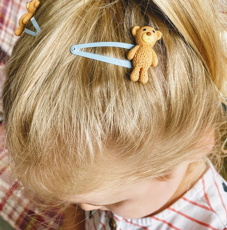 May include: A young girl with blonde hair wearing a red and white striped shirt has two blue hair clips in her hair. Each clip has a brown teddy bear charm.