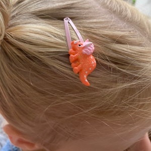 May include: A light pink metal snap hair clip featuring a textured orange triceratops dinosaur figurine with a pink frill and small white polka dot details on its body, securely attached to the surface of the hair accessory.