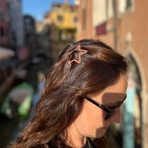 Pu&ograve; includere: Un fermaglio per capelli a forma di stella color rame &egrave; posizionato tra i capelli castani. Il fermaglio &egrave; un accessorio semplice ed elegante. Lo sfondo &egrave; una vista sfocata di edifici e un canale, che suggerisce un ambiente esterno.