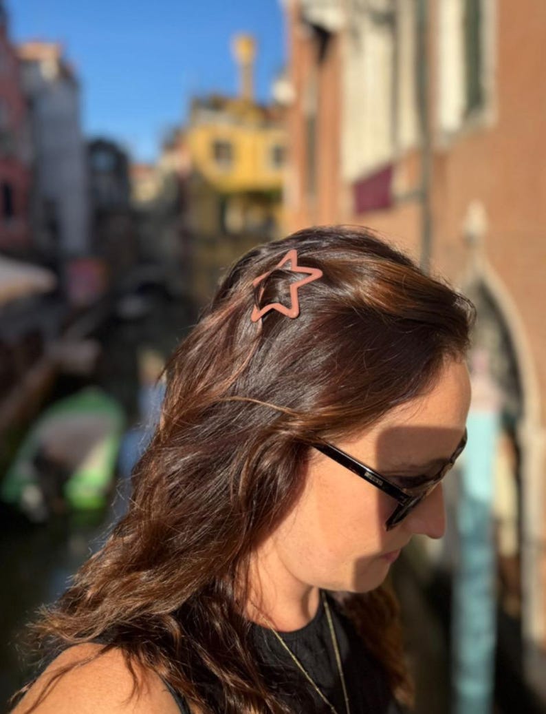 Pu&ograve; includere: Un fermaglio per capelli a forma di stella color rame &egrave; posizionato tra i capelli castani. Il fermaglio &egrave; un accessorio semplice ed elegante. Lo sfondo &egrave; una vista sfocata di edifici e un canale, che suggerisce un ambiente esterno.