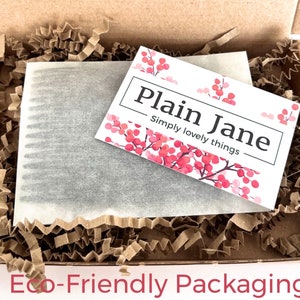 May include: A brown cardboard box filled with brown shredded paper and a white card with the text "Plain Jane Simply lovely things" and pink floral accents. The box contains a wrapped item.