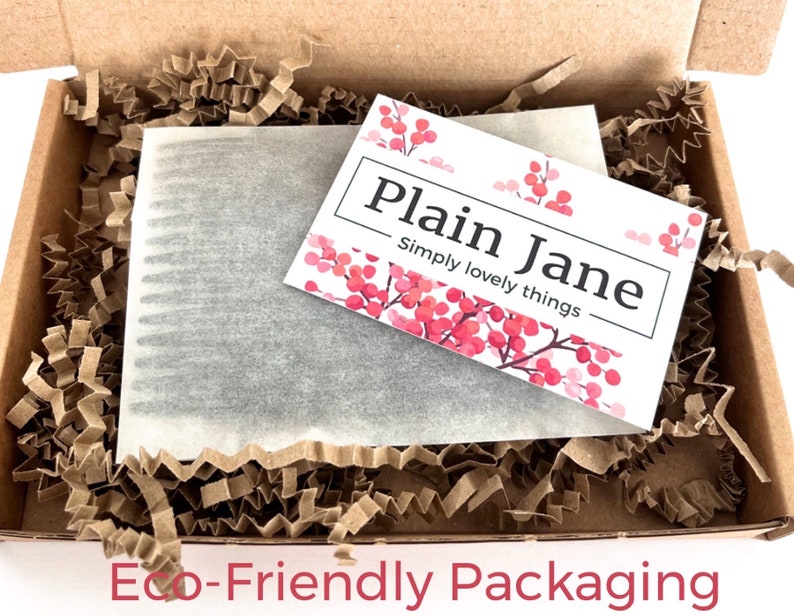 May include: A brown cardboard box filled with brown shredded paper and a white card with the text "Plain Jane Simply lovely things" and pink floral accents. The box contains a wrapped item.