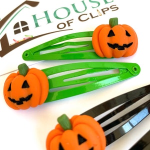 Op de afbeelding: Drie groene en zwarte haarclips met oranje pompoen-vormige bedels. De pompoenen hebben een jack-o'-lantern gezicht met zwarte ogen, een neus en een mond.