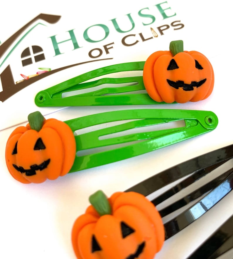 Op de afbeelding: Drie groene en zwarte haarclips met oranje pompoen-vormige bedels. De pompoenen hebben een jack-o'-lantern gezicht met zwarte ogen, een neus en een mond.