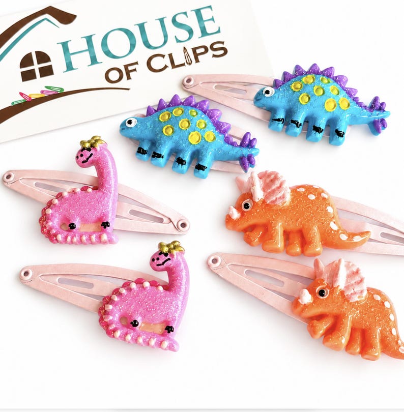 May include: Five hair clips featuring glittery dinosaur figures in blue, pink, and orange, attached to light pink metal snap clips. A white card with the text HOUSE OF CLIPS is visible in the background, displaying a logo of a house shape.