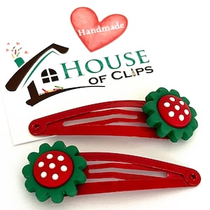 May include: Two red hair clips with green flower-shaped embellishments. Each flower has a red centre with white polka dots. The clips are labelled "House of Clips" and "Handmade".