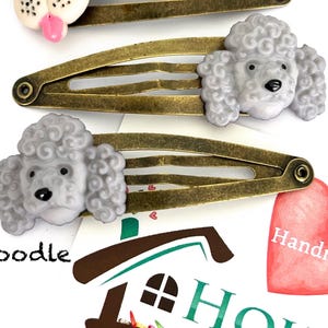 May include: Three hair clips with poodle designs. The clips have a bronze finish and feature gray poodle heads. One clip has a white dog head with a pink tongue. The word "Poodle" is printed on the paper background.