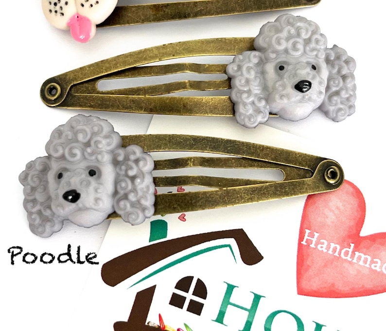 May include: Three hair clips with poodle designs. The clips have a bronze finish and feature gray poodle heads. One clip has a white dog head with a pink tongue. The word "Poodle" is printed on the paper background.