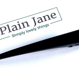 May include: A sleek, black hair clip with a pointed design. The clip is positioned next to a white card with the text "Plain Jane" and "Simply lovely things" in a green box.