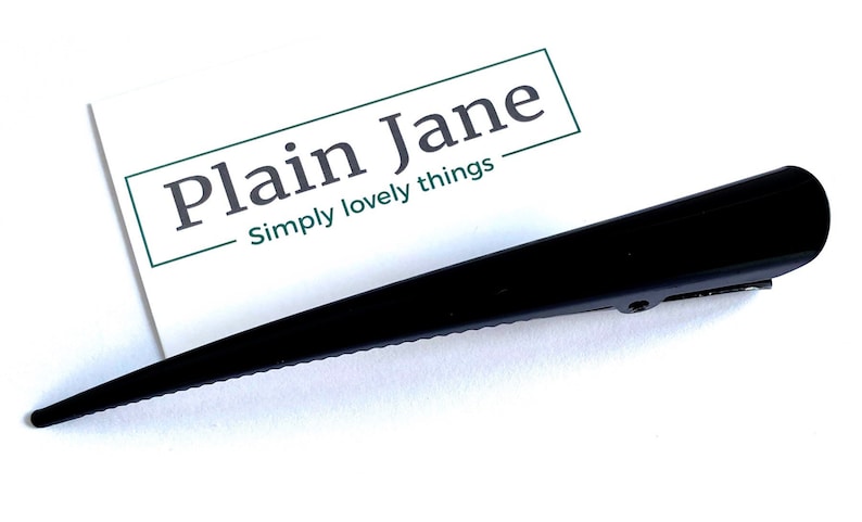 May include: A sleek, black hair clip with a pointed design. The clip is positioned next to a white card with the text "Plain Jane" and "Simply lovely things" in a green box.