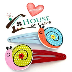 Può includere: Due fermagli decorativi con disegni di lumache. Uno è azzurro, l'altro rosso. I disegni delle lumache sono in legno e presentano pois e facce sorridenti. L'immagine include anche il testo "HOUSE OF CLIPS" e un cuore con la scritta "Handmade".
