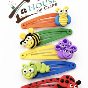 May include: Five metal hair clips featuring colorful clay insect figures: a blue and yellow bee, a yellow and black bee, a purple butterfly, a light green caterpillar, and a red and black ladybug. The logo House of Clips is visible.