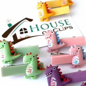 May include: A collection of colorful dinosaur-shaped hair clips. The clips feature various colors, including green, pink, yellow, and purple, with red and blue accents. The text "HOUSE OF CLIPS" is visible in the background.