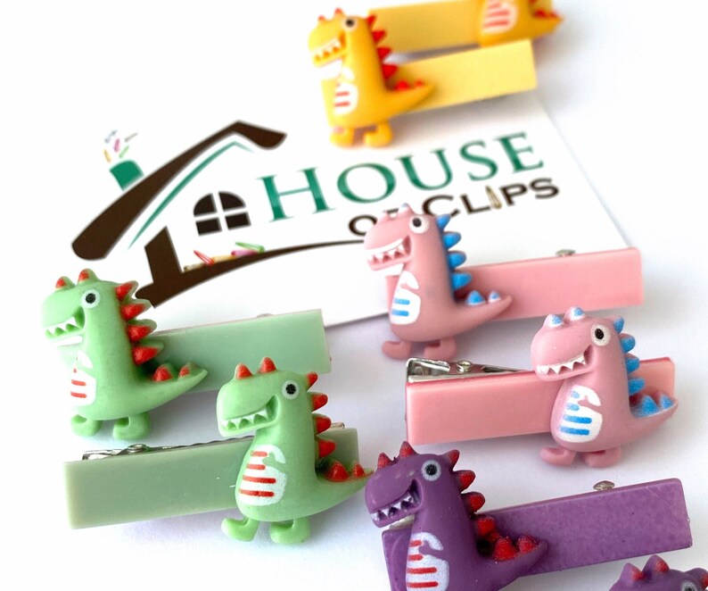 May include: A collection of colorful dinosaur-shaped hair clips. The clips feature various colors, including green, pink, yellow, and purple, with red and blue accents. The text "HOUSE OF CLIPS" is visible in the background.