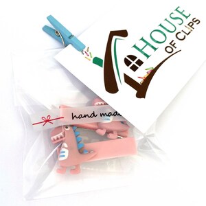 May include: A clear plastic bag contains two pink dinosaur hair clips with blue accents and a light blue clothespin. A white card with the text "HOUSE OF CLIPS" and a "hand made" sticker are also visible.