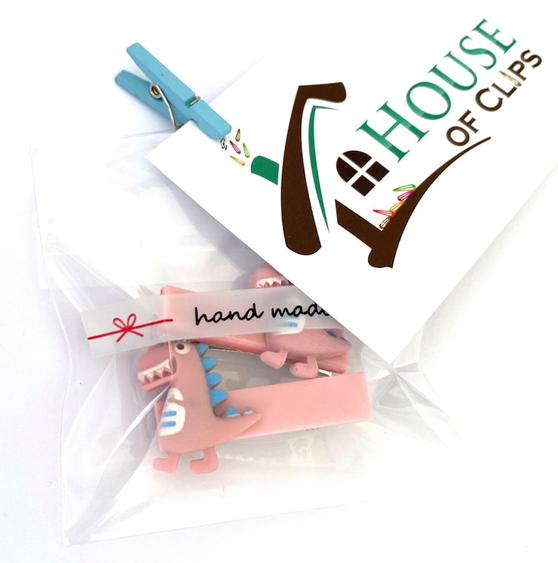May include: A clear plastic bag contains two pink dinosaur hair clips with blue accents and a light blue clothespin. A white card with the text "HOUSE OF CLIPS" and a "hand made" sticker are also visible.
