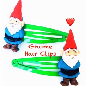 May include: Two green alligator hair clips with a gnome charm on each. The gnomes are wearing blue shirts, brown pants, and red hats. The text "Gnome Hair Clips" is written in red letters on the clips.